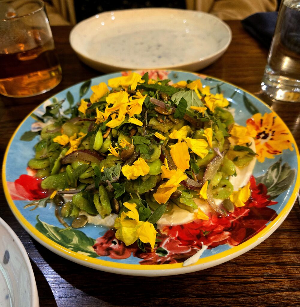 Fava bean salad at Cocina Consuelo on top of a teal plate with multicolored flowers. The fava bean salad is decorated with herbs, red onions and yellow edible flowers. The salad sits atop a white-tannish cashew cream.